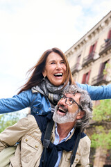 Vertical. Smiling elderly couple having fun together on vacation. Older man piggybacking mature woman enjoying leisure time visiting European city. Concept of excited people retirement relationships