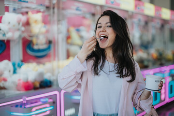 A smiling young woman enjoys ice cream against the backdrop of a vibrant amusement arcade full of plush toys.