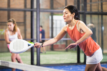 Portrait of concentrated asian woman playing paddle tennis indoors, preparing to hit forehand to...