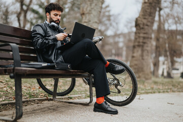 A young male entrepreneur in casual business attire is focused on his work outdoors, with a bike propped beside him on a park bench.