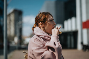 A young professional woman in a casual pink jacket eats a snack while standing in an urban setting...