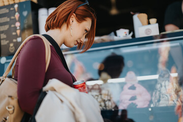 A thoughtful young woman with auburn hair gazes at delicious pastries while holding a drink and a tote bag at a bakery.