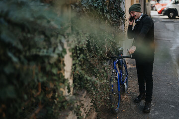 A fashionably dressed man talks on his smart phone while leaning on his bike, urban commuting scene with foliage.