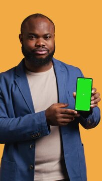 Vertical video Upset african american man showing thumbs down sign gesturing holding green screen phone, disapproving. BIPOC person doing rejection hand gesture regarding smartphone, studio background