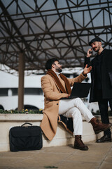 Capture of two professional men in fashionable attire having a discussion outside. One sits with a laptop while the other talks on the phone.