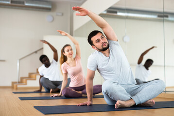 Man doing exercises in lotus pose with group, yoga training in gym.