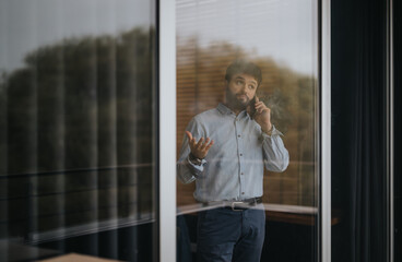 A professional businessman engaged in a conversation over the phone beside a modern office window, exuding confidence and success.