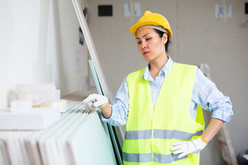 Woman in hardhat and yellow vest counting drywall sheets - obrazy, fototapety, plakaty