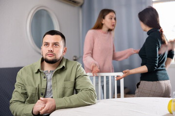 Upset man sitting at table while two women quarrel behind him