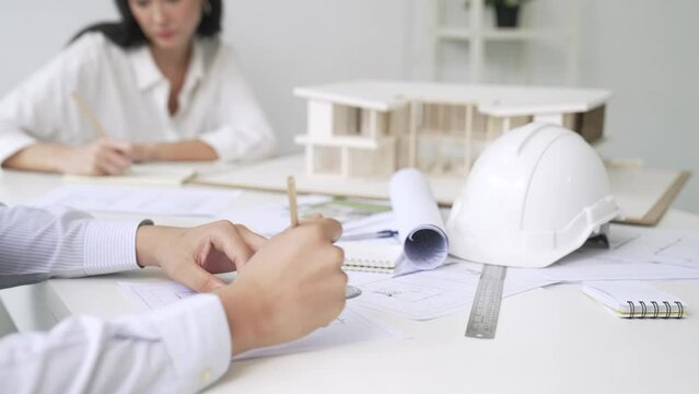 Professional architect engineer drawing blueprint and working together with young beautiful caucasian coworker on meeting table with safety helmet, house model, blueprint scatter around. Immaculate.
