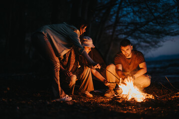 A joyful gathering of friends sharing stories around a warm campfire while preparing food outdoors by the lake.