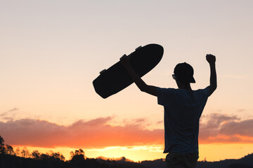 Silhouette of skater man celebrating with his longboard skate, with sunset sky in the background. 