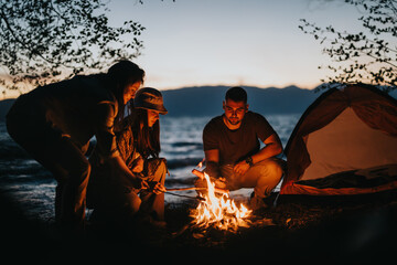 Happy friends gather around a campfire, preparing food during a camping trip by a serene lake at dusk.
