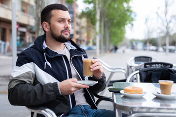 Young bearded man traveler enjoying warm cocoa with sweet soft donut at table of street cafe while...