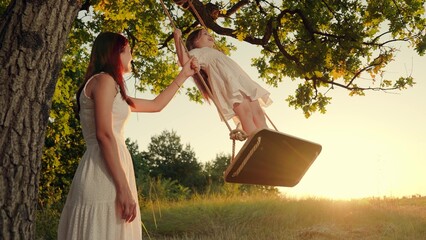 Little daughter Mother, swing on swing in park under tree at sunset. Happy family. Child, mother plays on wooden swing, dreams of flying. Family happiness, dreams, entertainment Concept. Baby swing