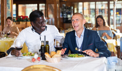 Two cheerful men friends enjoying evening meal with wine in restaurant