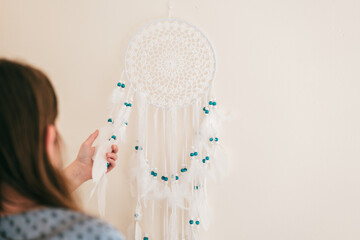 Dream Catcher.girl hangs a dream catcher on the wall.dream catcher with white feathers and blue beads on a beige wall.Decoration for home and room. Macrame wall decoration  - 788826317