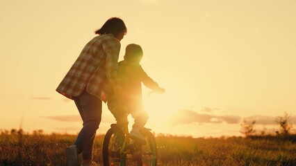Boy, his mother learn to ride bicycle in meadow in sun. Mom teaches her son to ride bike in park at sunset. Family day out in nature. Mother little son learn to ride bike together in summer. Teamwork