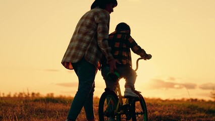 Mom teaches her son to ride bike in park at sunset. Family day out in nature. Mother little son learn to ride bike together in summer. Boy, his mother learn to ride bicycle in meadow in sun. Teamwork