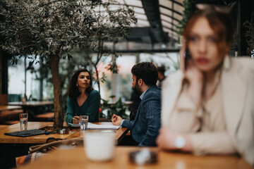 Two business professionals engage in a focused discussion over documents in a modern cafe environment, demonstrating teamwork and strategy. Blurred businesswoman having phone call. Copy space.