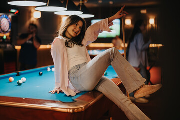 Carefree young woman sitting on a pool table, posing joyfully in a lively bar setting with friends...