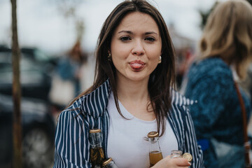Capturing a joyful moment at an outdoor event, a young woman playfully sticks out her tongue, holding soda bottles amidst a social setting.