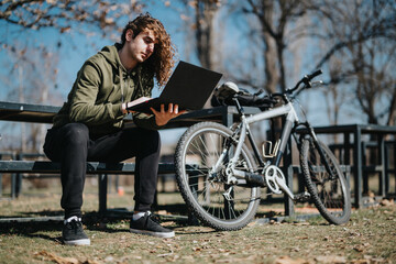 Handsome young man sits on a park bench with a laptop, relaxing near his bicycle on a sunny weekend day. Ideal for themes of leisure, technology in nature, and modern lifestyle.