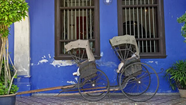 Two traditional hand pulled rickshaws parked together outside of an old colorful blue building in George Town, Malaysia