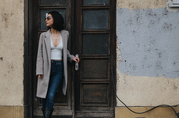 Elegant young woman leaning against an old textured door outdoors. She exudes confidence and style with her chic outfit and sunglasses.