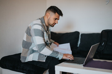 Focused young businessman reviews paperwork while using his laptop seated on an ottoman in a well-lit room