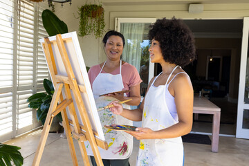 Biracial mother and adult daughter are painting together at home, both wearing aprons - 788839917