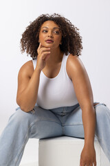 Fototapeta na wymiar Biracial young female plus size model sitting, touching chin, looking thoughtful on white background