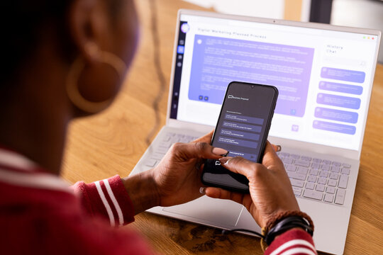 African American Young Woman Indoors With Phone, Laptop Showing AI Chat Bot Screen At Home