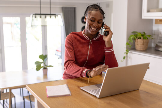 African American young woman talking on the phone indoors at home, looking at a laptop