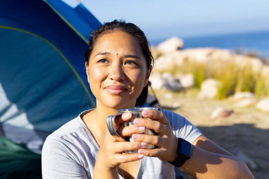 Biracial female hiker holding cup, sitting by tent
