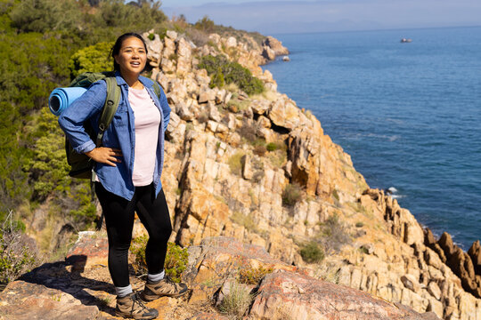 Biracial female hiker standing on rocky terrain, overlooking the sea, adventure in nature