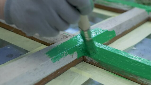 Gloved hand guides green paint onto wooden window. Applied with brush. Patch of old wooden surface, showing through cracked and flaking paint. Handiwork, restore woodwork outdoor