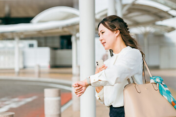 駅・ターミナル・バス停で腕時計で時間を確認する若いビジネスウーマン（遅刻・遅れる・焦る・イライラ）
