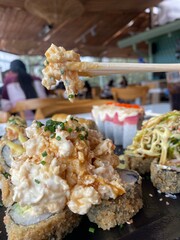 Chopsticks holding a piece of tempura shrimp from a Nikkei-style sushi board, with the blurred background of a restaurant