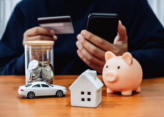 Monthly expenses, budget, financial insurance concepts. White home, car, money coin bottle and piggy bank on table while person holding credit card and using mobile phone for payment application.