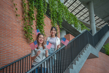 Two Asian female college students stand holding American flags. On the stairs in the university Look and smile happily.
