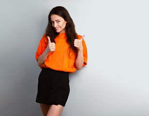 Happy toothy smiling beautiful woman showing thumb up sign by two hands in bright orange t-shirt and black skirt on blue studio wall background - 788891773