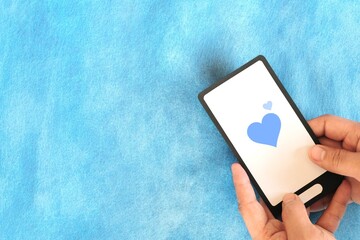 Human hand holding mobile phone with blue heart icon. Kindness, love and positivity in social media internet concept.