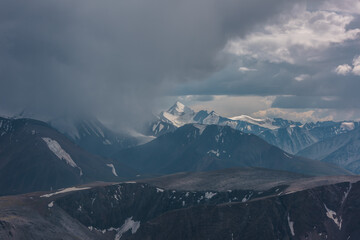 Dark atmospheric landscape with mountain silhouettes and large snow-capped peaked top in rainy low clouds. Dramatic alpine view to high mountains in gray cloudy sky. Big snowy mountain range in rain.
