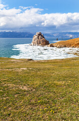 Spring on Baikal Lake. View of natural landmark - Shamanka Rock during ice drift from green lawn with yellow dandelions on the coast of Olkhon Island on sunny May day. Spring holidays and relaxation