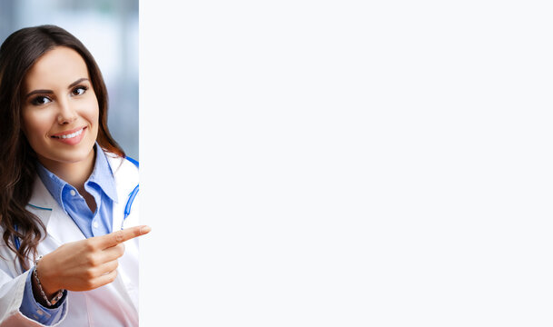 Portrait image of happy smiling female doctor pointing blank signboard with copy space empty area for ad slogan text. Young brunette beautiful woman in medical, clinic, healthcare concept