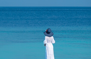 The woman wears a white Hawaiian dress and she wears a blue Hawaiian hat. She stands looking at the view of the Andaman Sea. Travel concept.