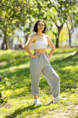A woman is standing in a park wearing a white tank top and grey sweatpants