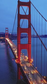 Vertical video. San Francisco landmark at sunset on cloudy day, aerial view of the Golden Gate Bridge and city skyline. Cinematic drone footage of red bridge with night illumination on blue sky