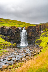 Cover page with copy space, Gufu waterfall with dramatic sky and green hills. Beautiful Icelandic landscape, huge cliff, water cascade and rock in moss